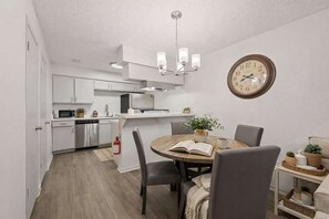 A modern kitchen with white cabinets, a microwave, and a sink. There's a dining table with chairs and a book, a clock on the wall, and a fire extinguisher on the floor. - Entertainment District Escape • 2BR + Parking (Arlington)