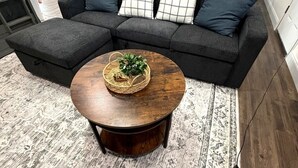 Une table basse ronde en bois, surmontée d’un panier tressé et d’une petite plante, est placée devant un canapé d’angle gris foncé agrémenté de coussins à motifs.