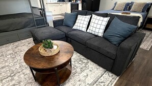 Un salon moderne avec un canapé d’angle gris foncé, une table basse ronde en bois surmontée d’un panier en osier et un tapis à motifs.