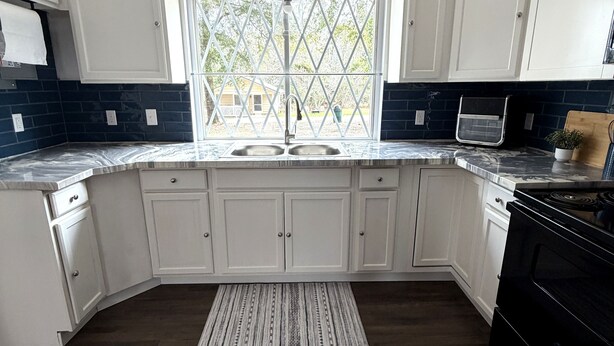 Une cuisine avec des meubles blancs, une cuisinière noire, une fenêtre donnant sur l’extérieur et un tapis à motifs.