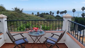 Ein Balkon mit zwei Klappstühlen und einem Tisch, der auf eine Küstenlandschaft mit Palmen und dem Meer blickt.
