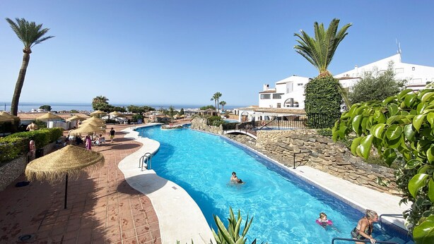 A swimming pool with a curved edge, surrounded by stone walls and palm trees, with a view of the sea and a white building in the background. - Las Mimosas 7B Capistrano Village in Nerja (Nerja)