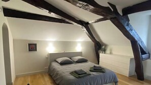 A bedroom with a sloped ceiling, exposed wooden beams, a bed with grey bedding, a bedside table, and a chair.