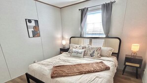 Ein ordentlich bezogenes Bett mit einer braunen Tagesdecke, ein Kopfteil mit einem Fenster und Vorhängen, zwei Nachttische mit Lampen und ein an der Wand hängendes Bild.