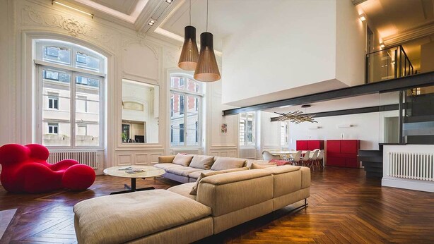 A spacious living room with a large beige sectional sofa, a red bean bag chair, and a round coffee table. There are multiple windows providing natural light, and a kitchen area is visible in the background. - LOFT · KEYWEEK contemporary Loft style Apartment in Biarritz City center (Biarritz)