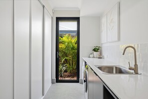 A narrow kitchen with a built-in washing machine, a stainless steel sink, and a door leading to a garden. - Coastal Luxe in Seaside Estate | 4 Bedrooms | Walk to Beach (Mandurah)