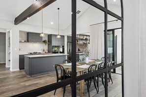 A modern kitchen with a central island, pendant lighting, and a dining area with a wooden table and chairs. - Coastal Luxe in Seaside Estate | 4 Bedrooms | Walk to Beach (Mandurah)