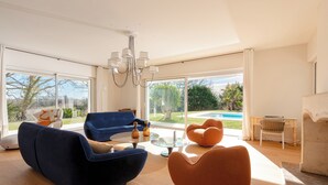 Een ruime woonkamer met een groot raam, een blauwe bank, een ronde oranje fauteuil en een glazen salontafel.