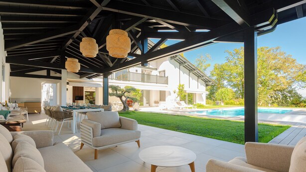 Een overdekt terras met banken en een tafel, een zwembad en een huis op de achtergrond.