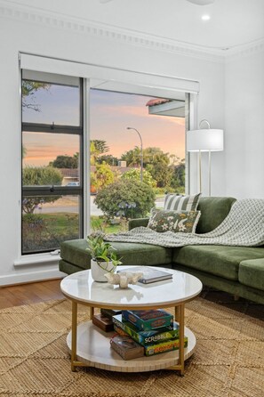Ein Wohnzimmer mit einem grünen Sofa, einem runden Couchtisch mit Gesellschaftsspielen, einem Fenster mit Blick auf Grünflächen und einen Sonnenuntergang.