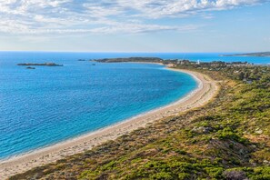 Eine Küstenlandschaft mit Sandstrand, klarem blauen Wasser und hügeligem Gelände mit spärlicher Vegetation.