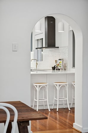 Eine moderne Küche mit weißen Schränken, einer schwarzen Dunstabzugshaube und einer weißen Spritzschutzwand. Es gibt drei weiße Barhocker und einen Holztisch.