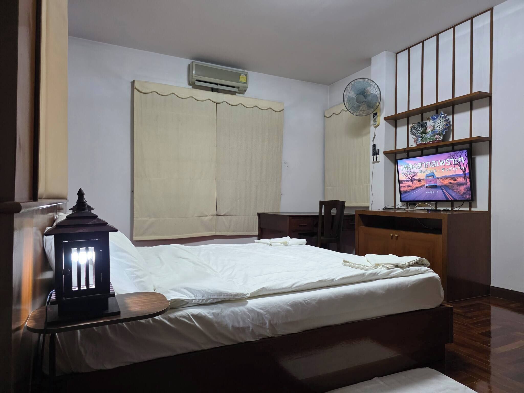 A bedroom with a bed, bedside table, lamp, air conditioner, fan, TV, and a wooden shelf.