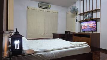 A bedroom with a bed, bedside table, lamp, air conditioner, fan, TV, and a wooden shelf.