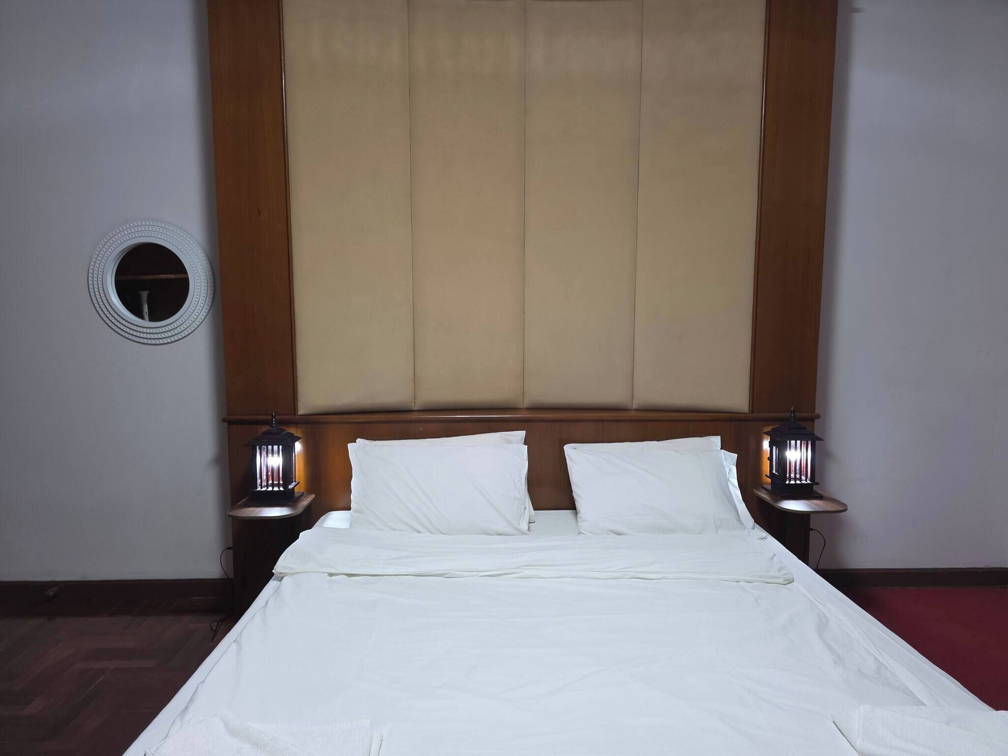 A hotel room with a large bed, two bedside tables with lamps, a round mirror on the wall, and a large window with a beige blind.