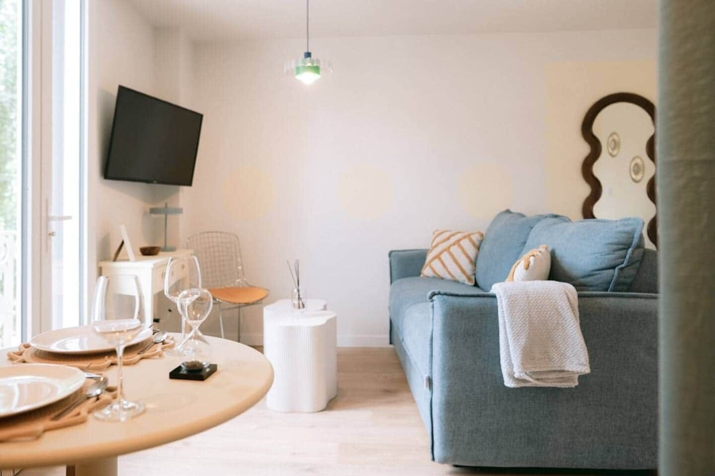 A living room with a round table set for dining, a grey sofa with a blanket, a white side table, and a mounted TV.