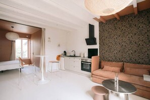 A modern living room with a sofa, a dining table, and chairs. There is a kitchen area with a stove and a sink. - Georges' Journey - 154 (Périgueux)