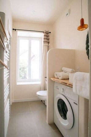 A compact bathroom with a washing machine, a toilet, and a window with curtains. - Georges' Journey - 154 (Périgueux)