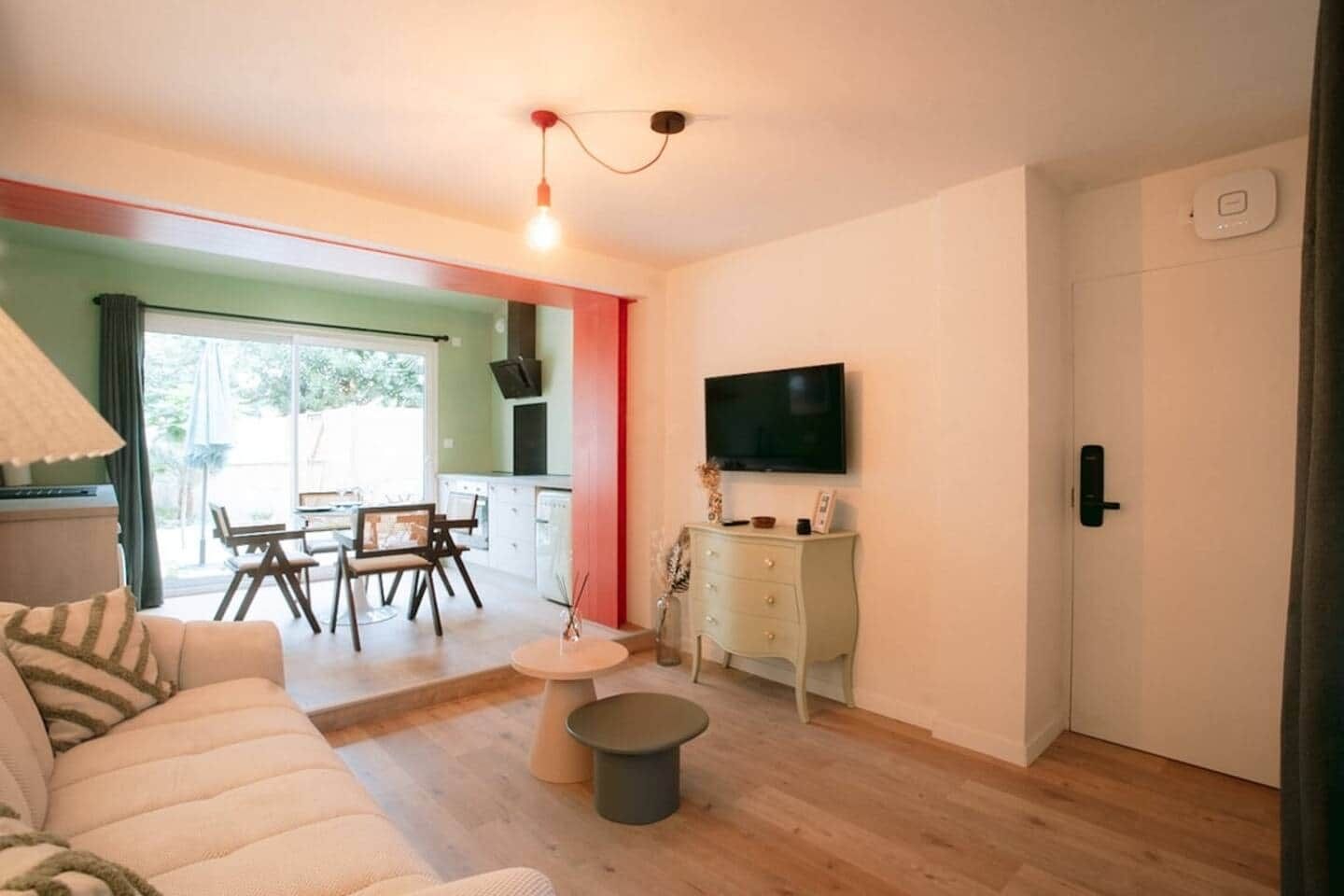Uma sala de estar com um sofá, uma mesa de jantar com cadeiras, uma TV fixada na parede e um pequeno armário.