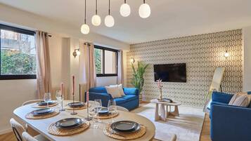 Uma área de refeições com uma mesa redonda posta para quatro pessoas, um sofá azul, uma pequena mesa lateral e uma TV montada na parede.
