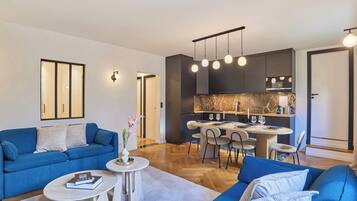 Uma sala de estar moderna com um sofá azul, uma área de jantar com mesa e cadeiras, e uma cozinha com armários escuros e painel de parede em mármore.