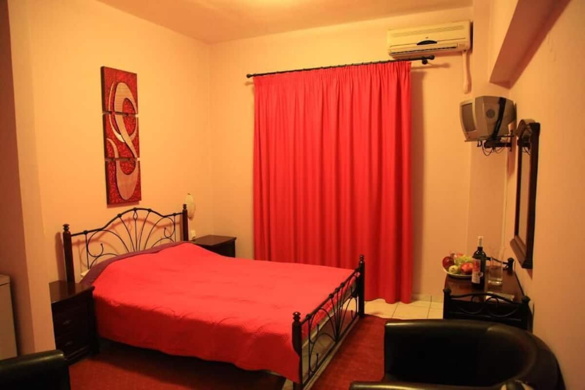 Een slaapkamer met een bed, nachtkastje, stoel, bureau met lamp, rood gordijn en een wandairconditioner.