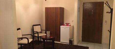 Une petite pièce agréable, avec un tapis marron foncé, une armoire en bois, un réfrigérateur blanc et une porte dotée d’un porte-manteau.