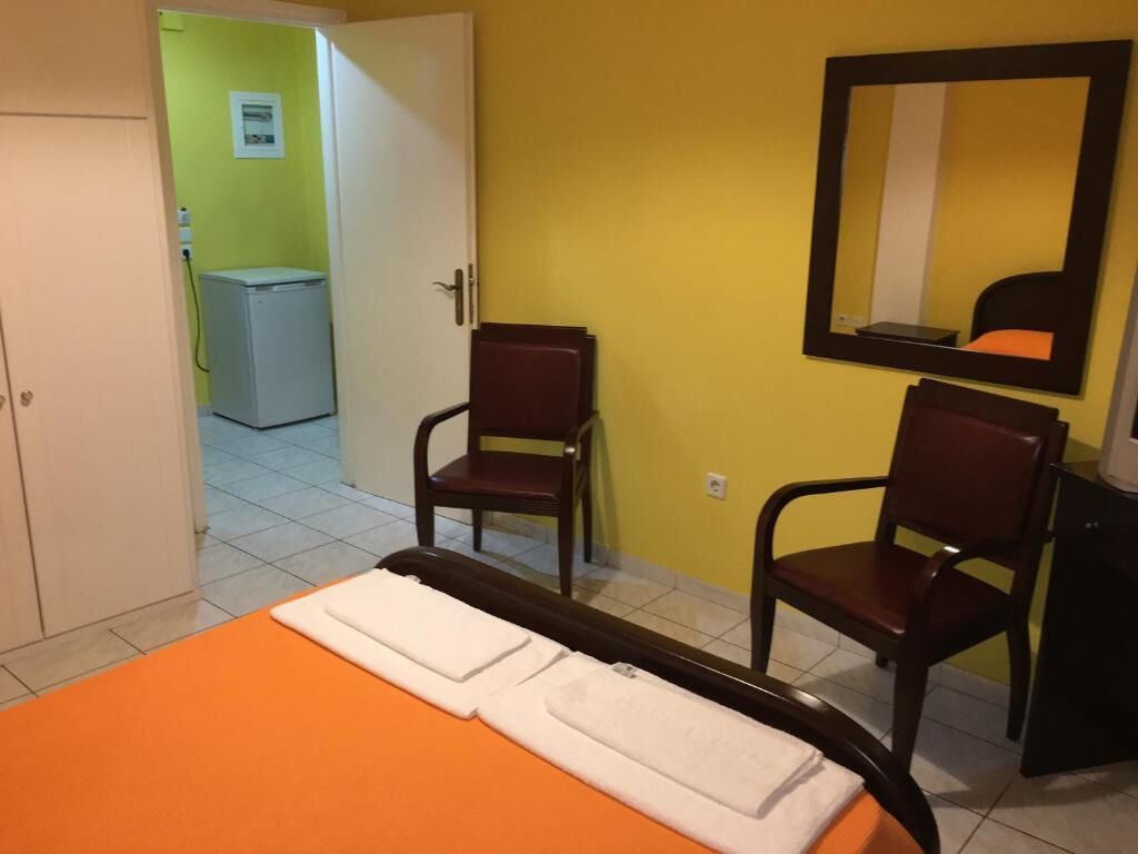 Une chambre d’hôtel avec un lit orange, deux chaises en bois, un miroir et un petit bureau.