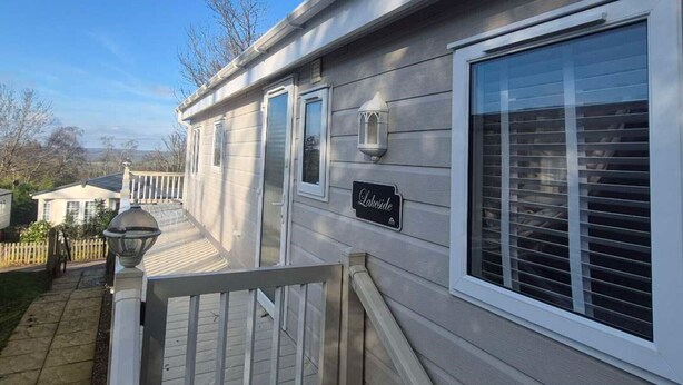 Une maison mobile dotée d’une véranda, d’une enseigne indiquant « Au bord du lac » et d’une fenêtre avec des stores.
