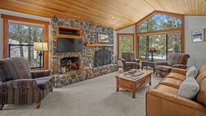A cozy living room with a stone fireplace, wooden ceiling, and large windows offering a view of trees. - Quiet Sunriver Retreat with Access to Exclusive Golf (Sunriver)