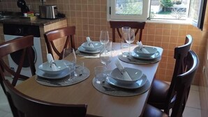 A dining table set with plates, glasses, and cutlery, surrounded by wooden chairs in a kitchen with a window and tiled backsplash. - Vionta: Your cozy retreat with garden view near the beach (Aguiño)