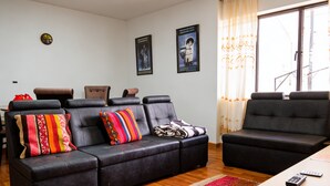 A living room with a sofa, armchairs, a coffee table, and a TV. There are posters on the wall and a window with curtains.
