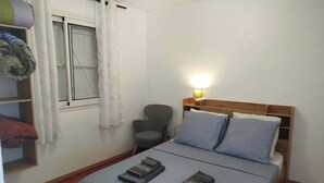 Un dormitorio con cama, una silla, una mesita de noche con lámpara y una ventana con cortinas.