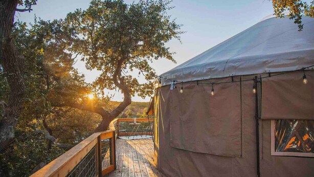 A wooden deck leading to a large tent with string lights, surrounded by trees and a clear sky. - Yurt in the trees w TX stars & Pickleball (Dripping Springs)