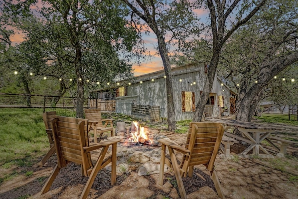 A rustic outdoor seating area with wooden chairs and a fire pit, surrounded by trees and a building with string lights. - Live Oak Lodge ↠ Rustic Barndo w Dog (or Kid) Run, Pickleball (Dripping Springs)