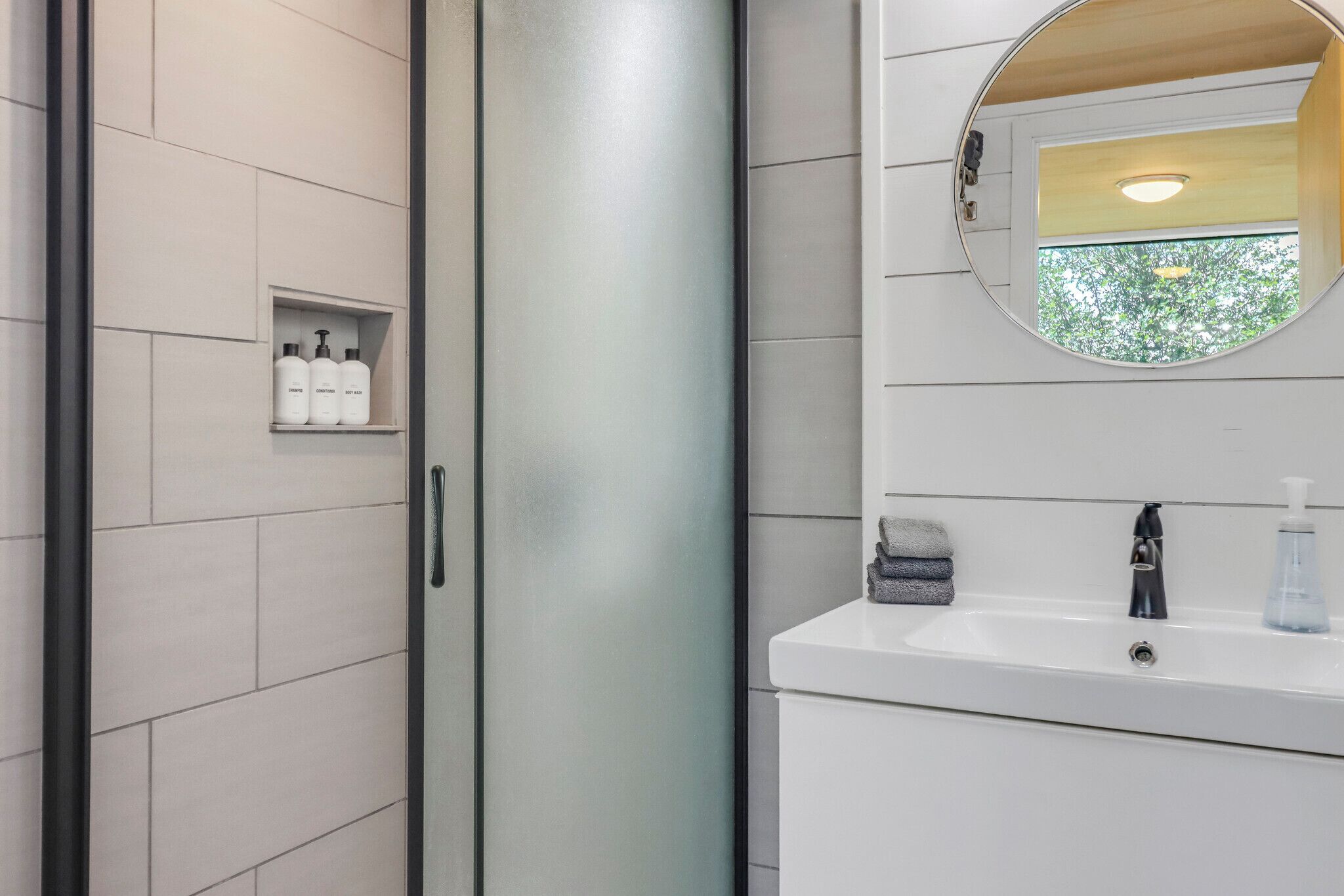 A modern bathroom with a glass shower enclosure, a white sink with a mirror above it, and a shelf with toiletries.