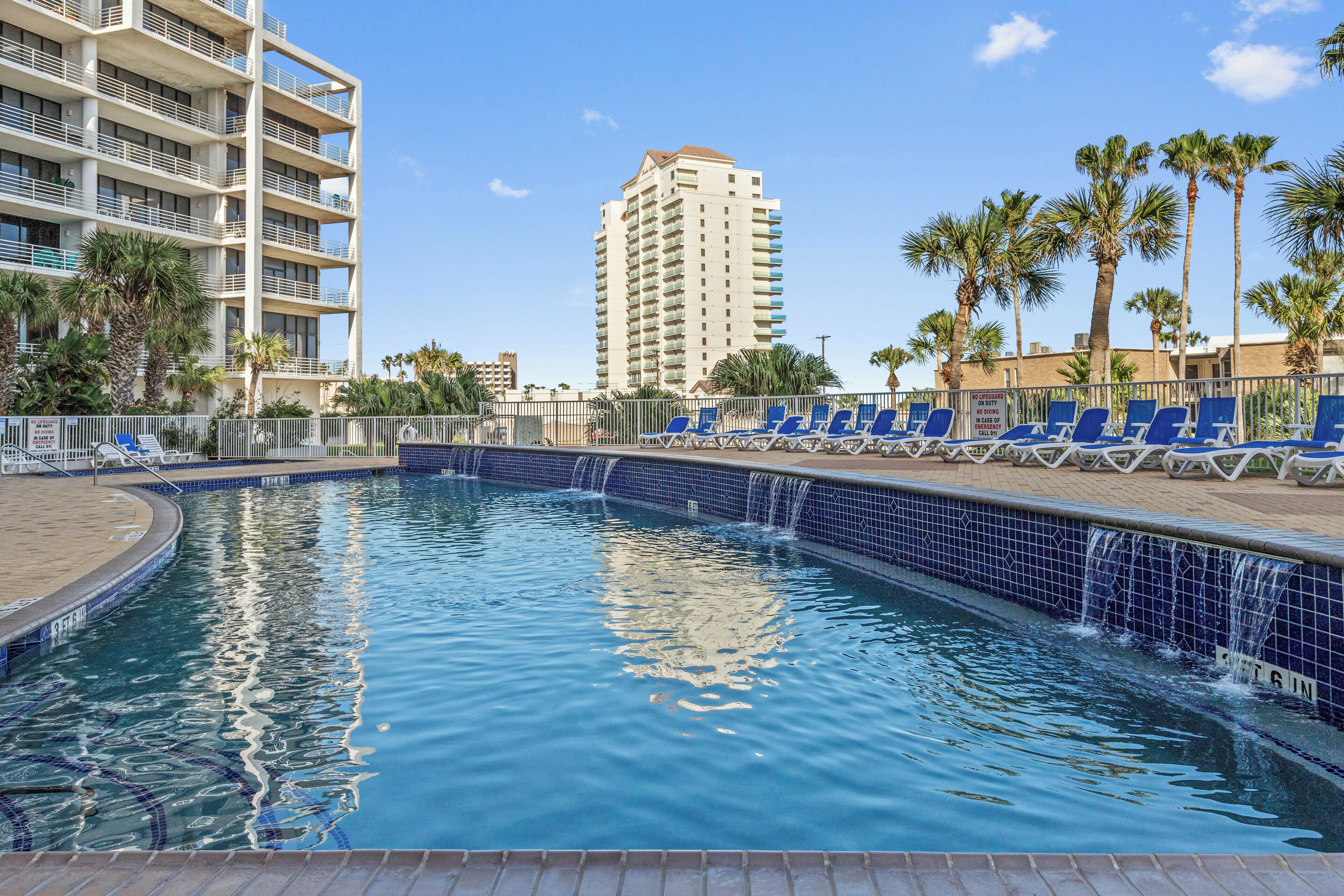 Una piscina con piastrelle blu, lettini da relax, palme e un edificio alto sullo sfondo.