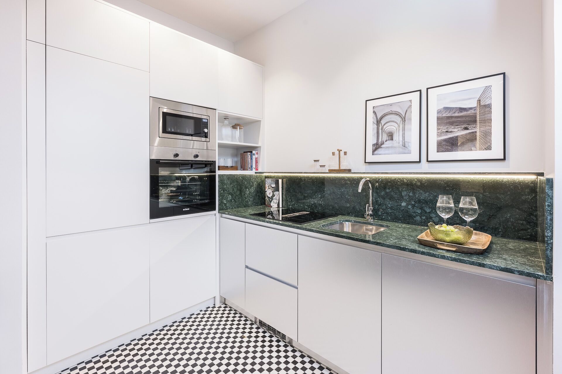 Een moderne keuken met witte kastjes, een zwart-wit geruite vloer, een ingebouwde oven, een gootsteen met een marmeren aanrechtblad en twee ingelijste foto's aan de muur.