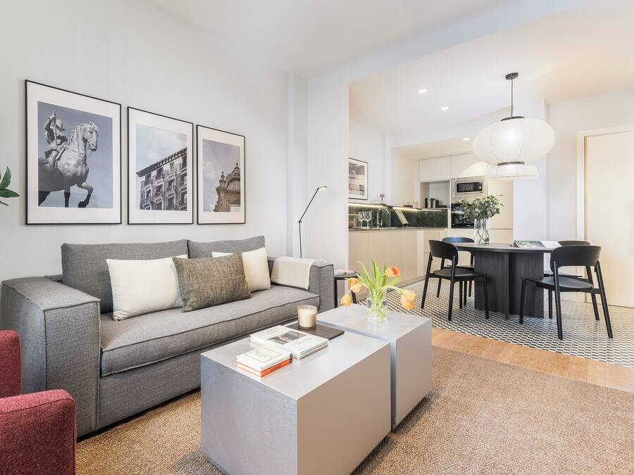 Una sala de estar moderna con un sofá gris, un sillón rojo y una mesa de centro con libros y un florero. Se observa un comedor con una mesa y sillas, y una cocina al fondo.