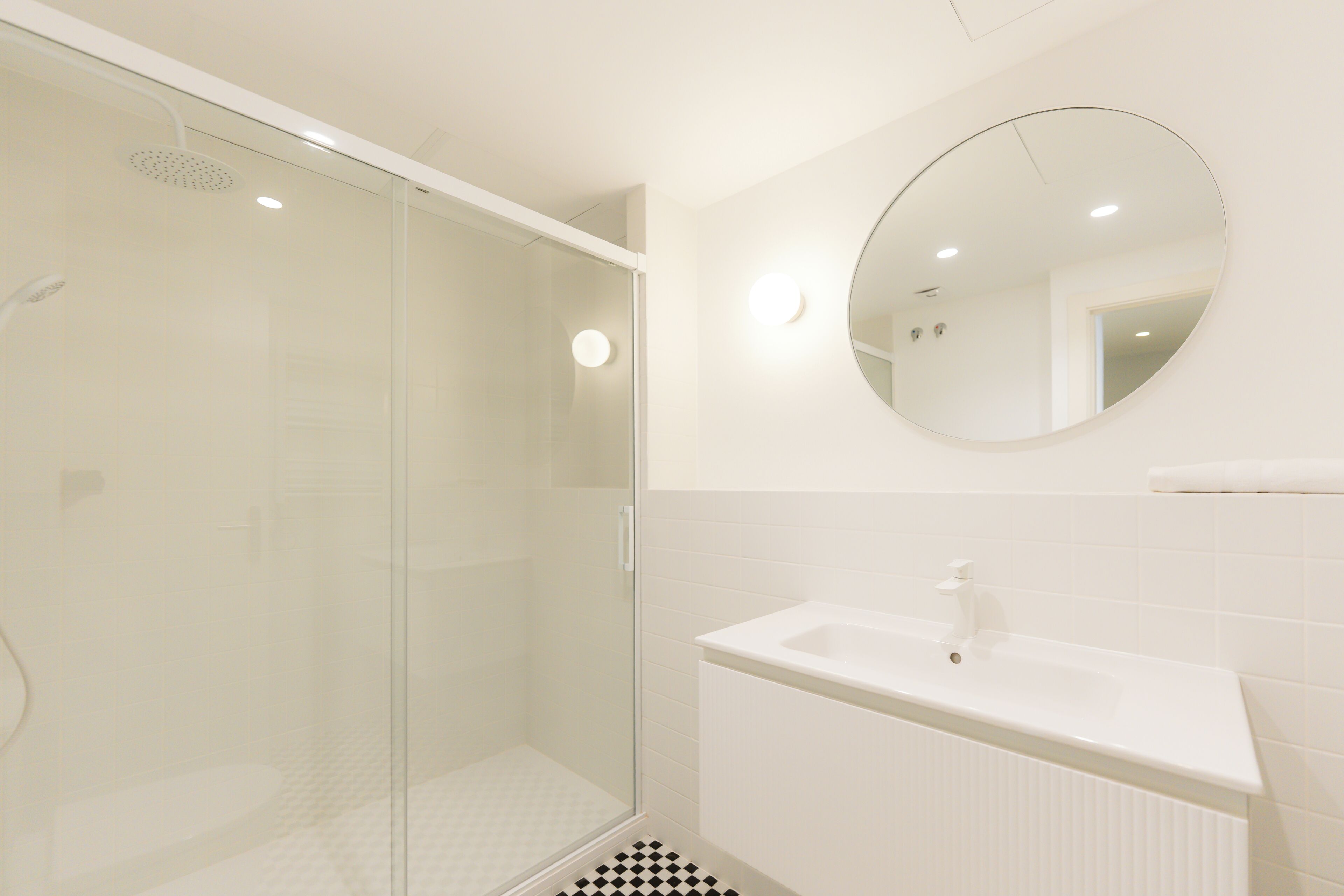 Un baño moderno con cabina de ducha de vidrio, un lavabo blanco y un espejo redondo en la pared.