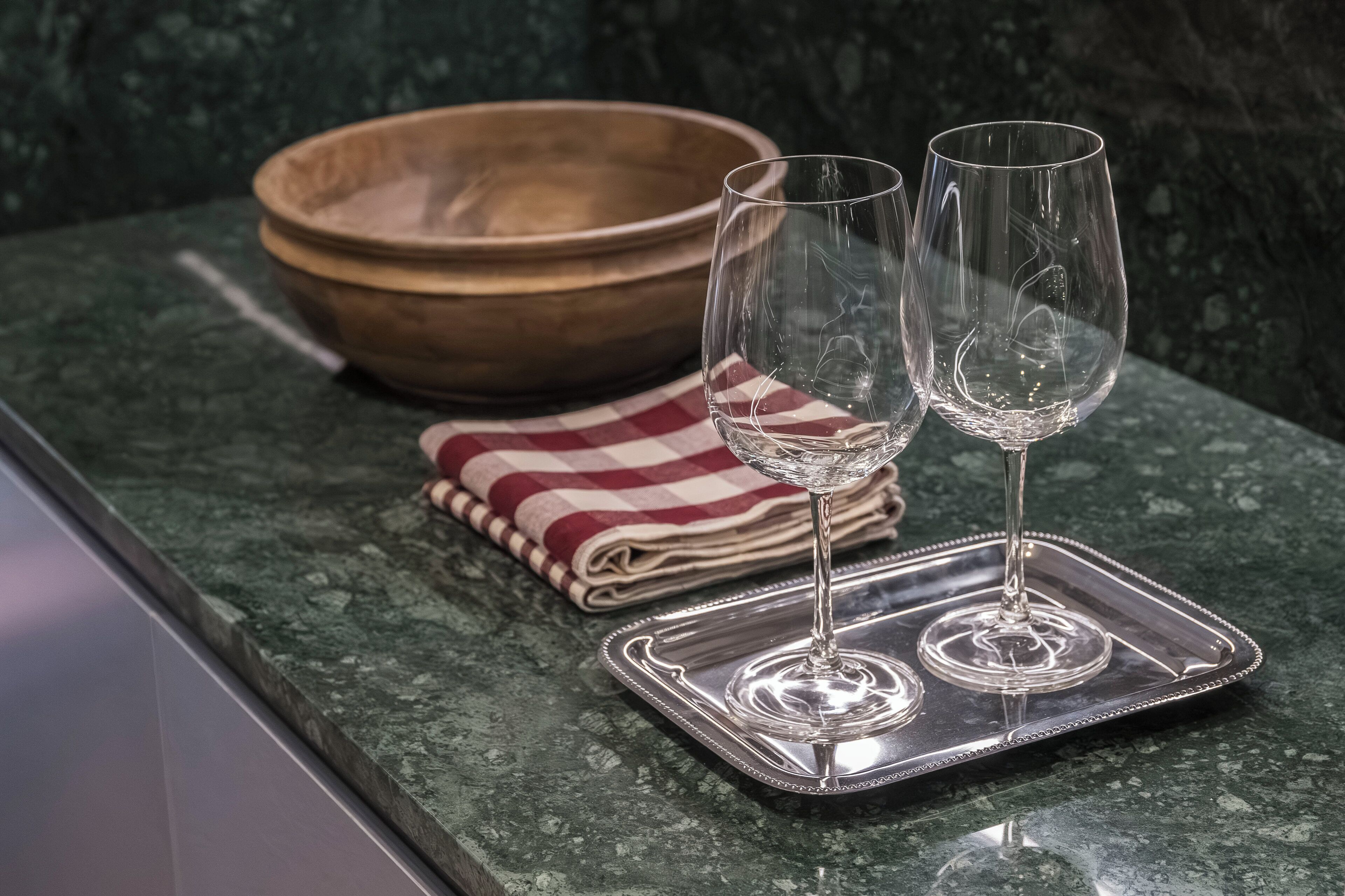 A kitchen counter with a wooden bowl, two wine glasses, and a folded red and white checkered cloth.