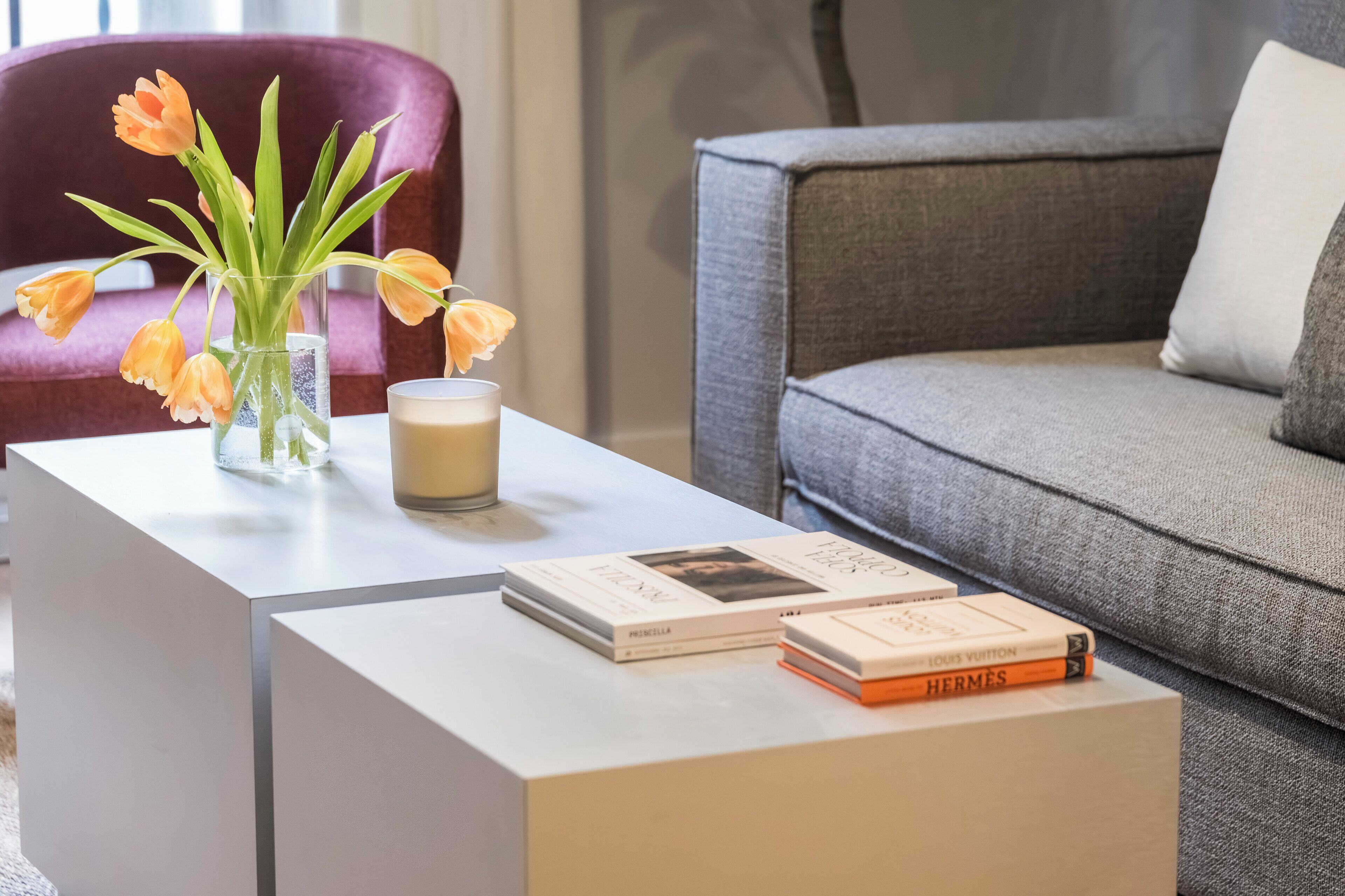 Un salón moderno con una mesa de centro blanca, un jarrón con tulipanes naranjas, una vela y dos libros.