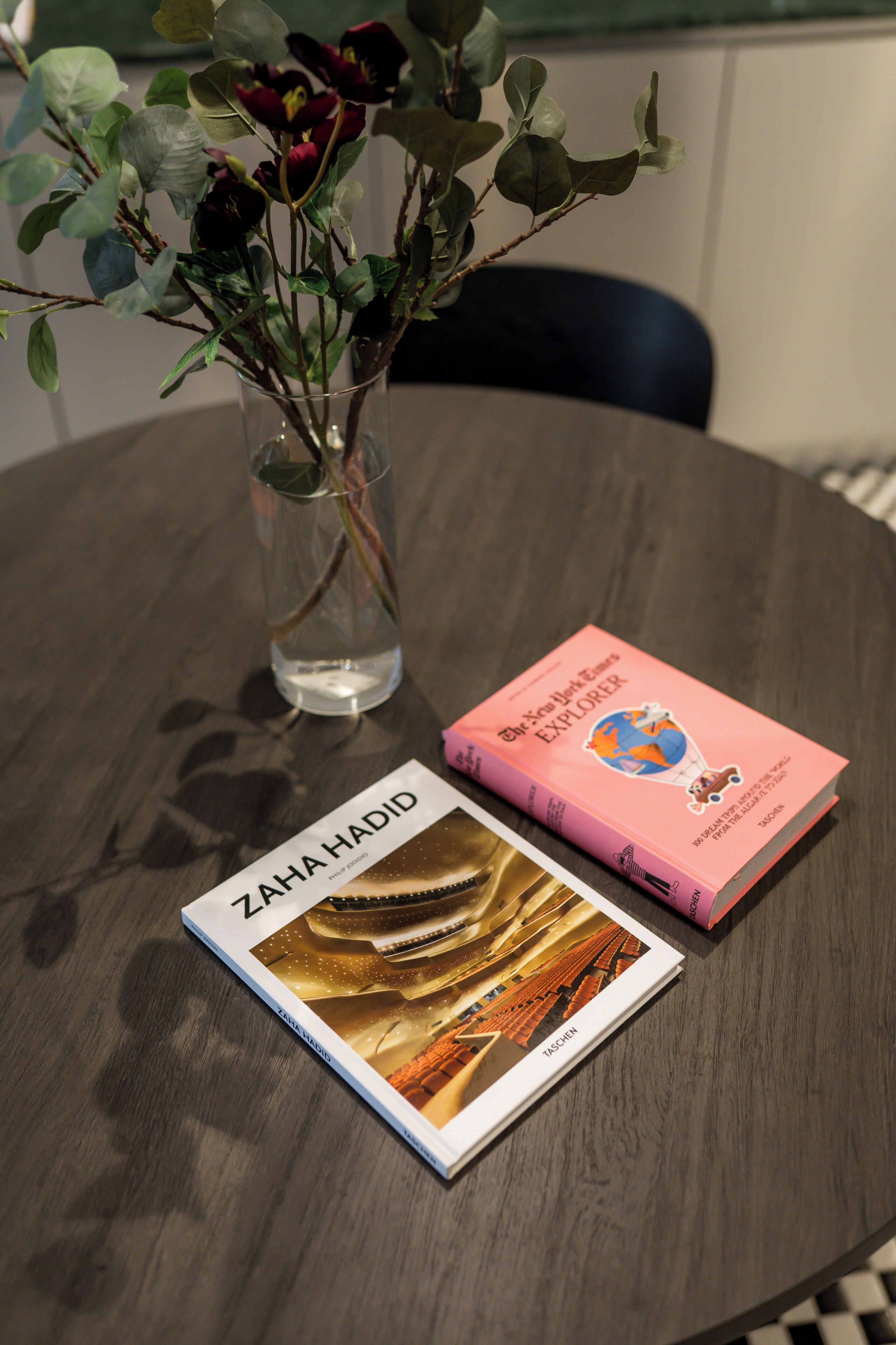 A book titled 'ZAHAVA HADID' and 'The New York Explorer' on a round table with a clear glass vase containing green leaves.