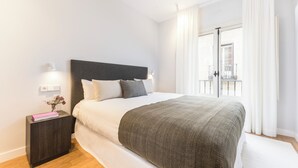 A neatly made bed with a dark headboard, white bedding, and a grey throw blanket. There is a small bedside table with a book and a vase of flowers. The room has white walls and a large window with curtains. - Edmond Vibes Plaza de España Suites (Madrid)