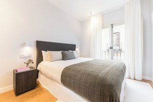 Ein ordentlich bezogenes Bett mit dunklem Kopfteil, weißer Bettwäsche und grauer Tagesdecke. Ein kleiner Nachttisch mit einem Buch und einer Blumenvase steht daneben. Der Raum hat weiße Wände und ein großes Fenster mit Vorhängen.