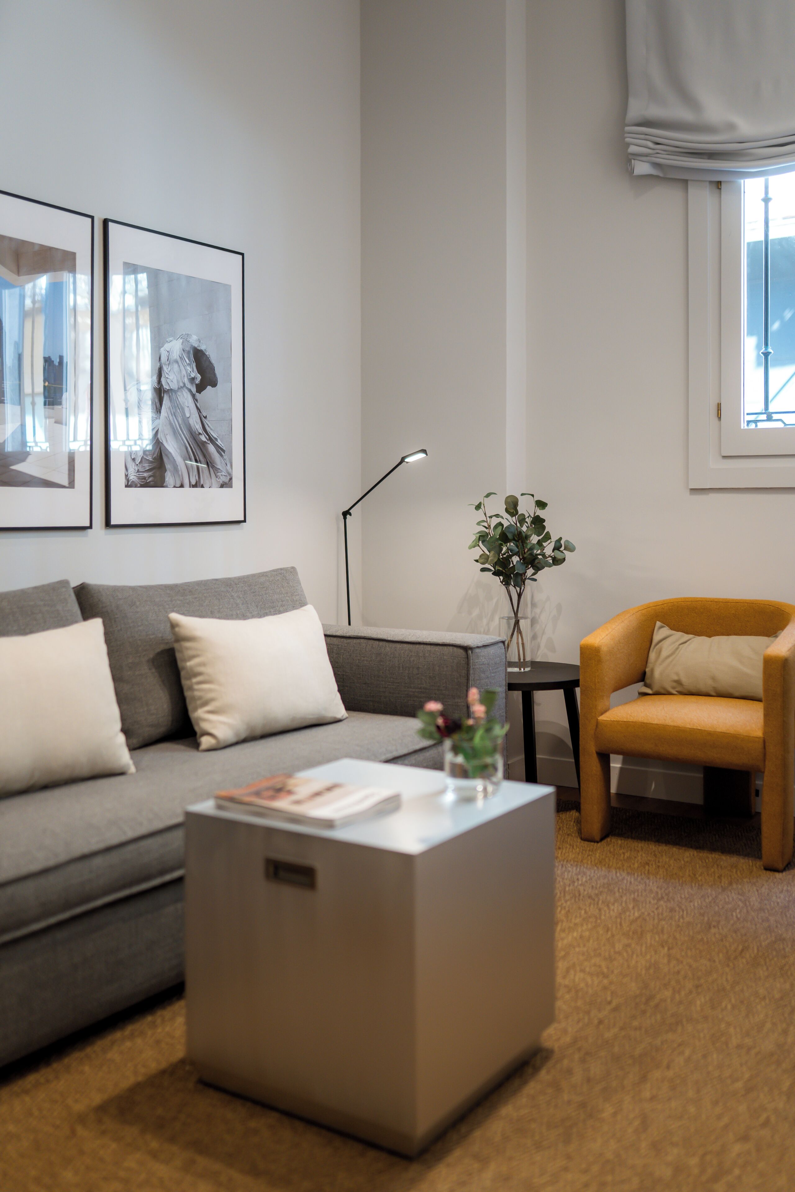 Un salon moderne avec un canapé gris, une petite table basse, un fauteuil jaune, une lampe sur pied et deux tableaux encadrés accrochés au mur.