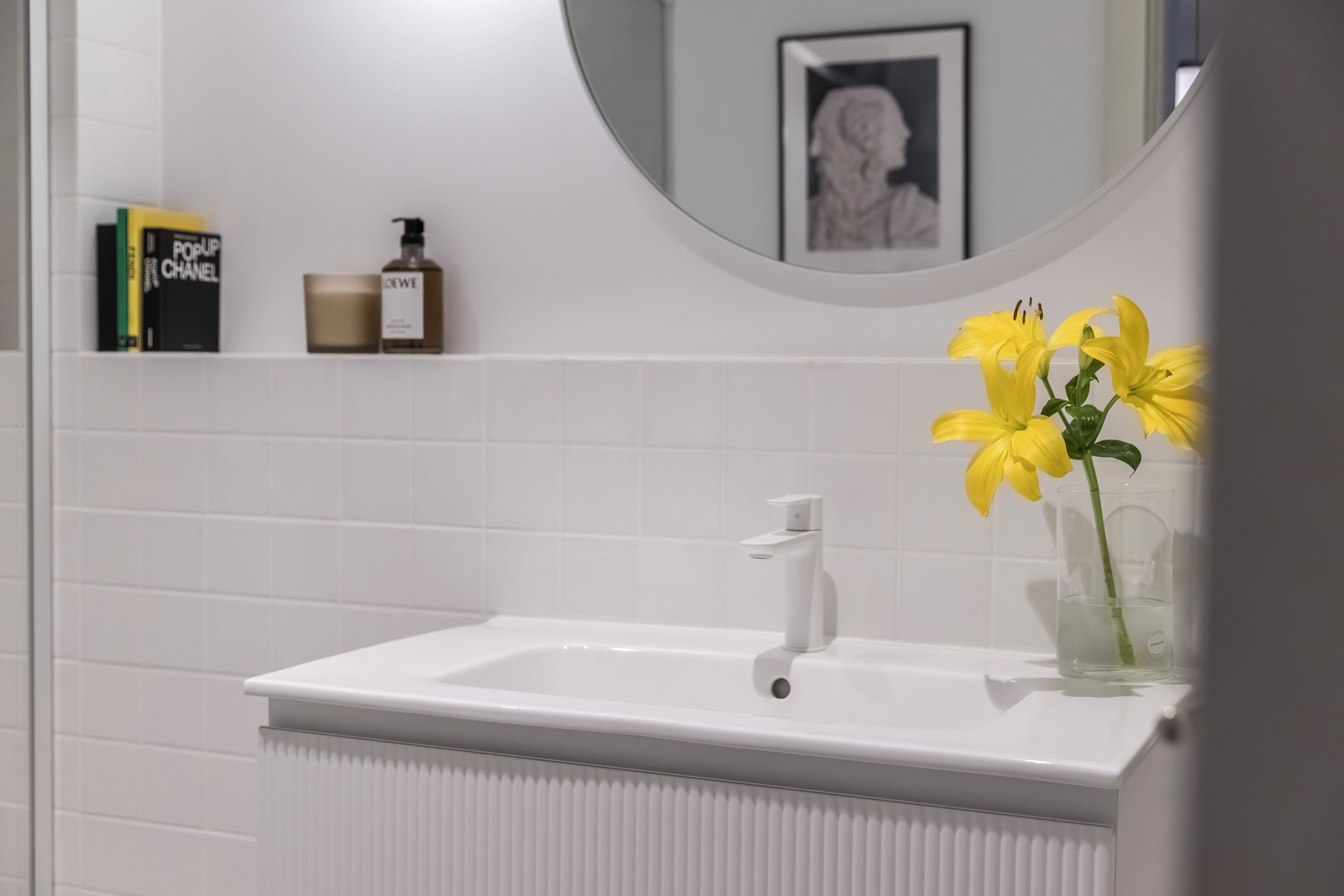 A bathroom with a white sink, a mirror, and a vase with yellow flowers. There are books and a bottle on the shelf.