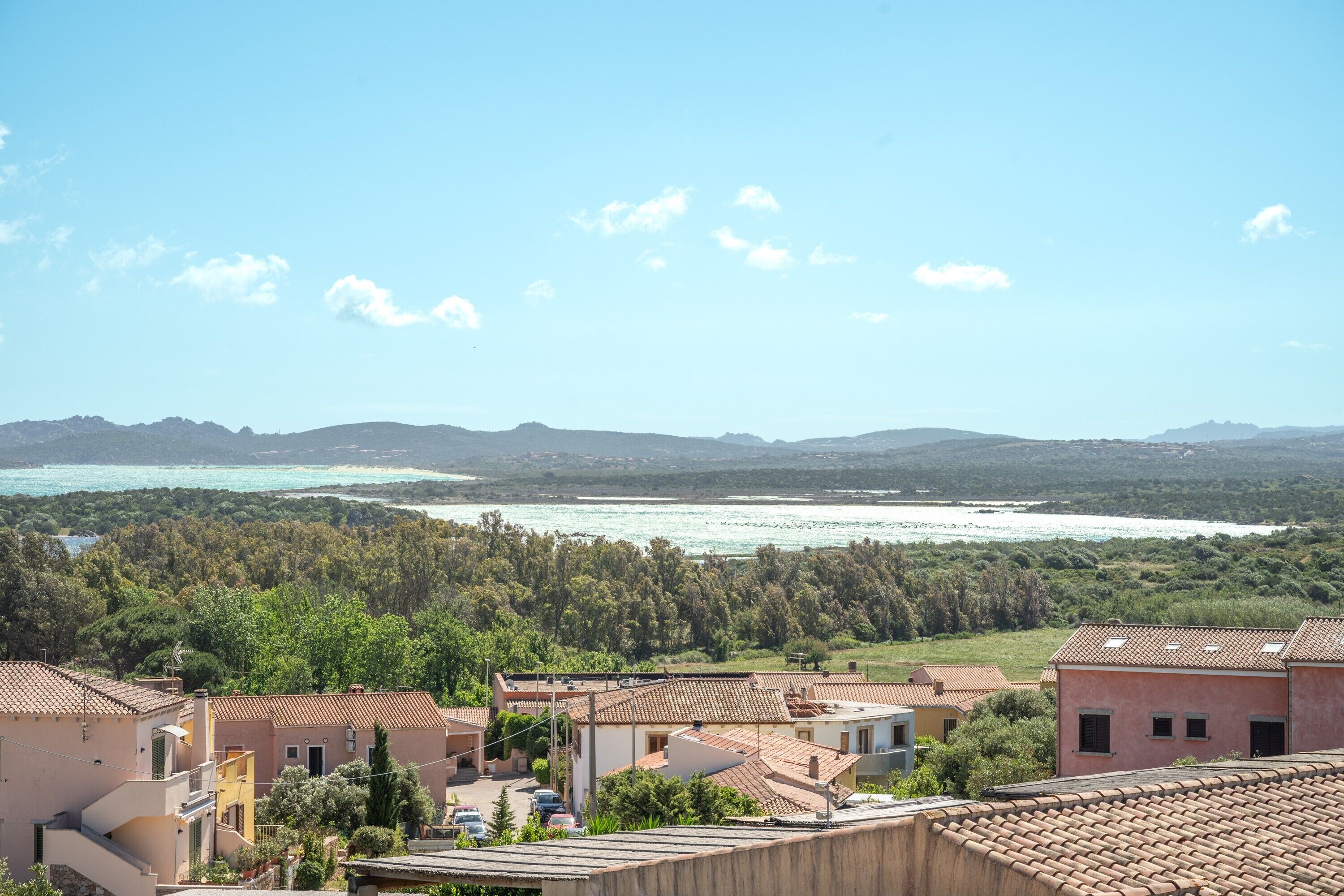 A coastal residential area with houses, a clear sky, and a view of the sea and mountains in the distance.