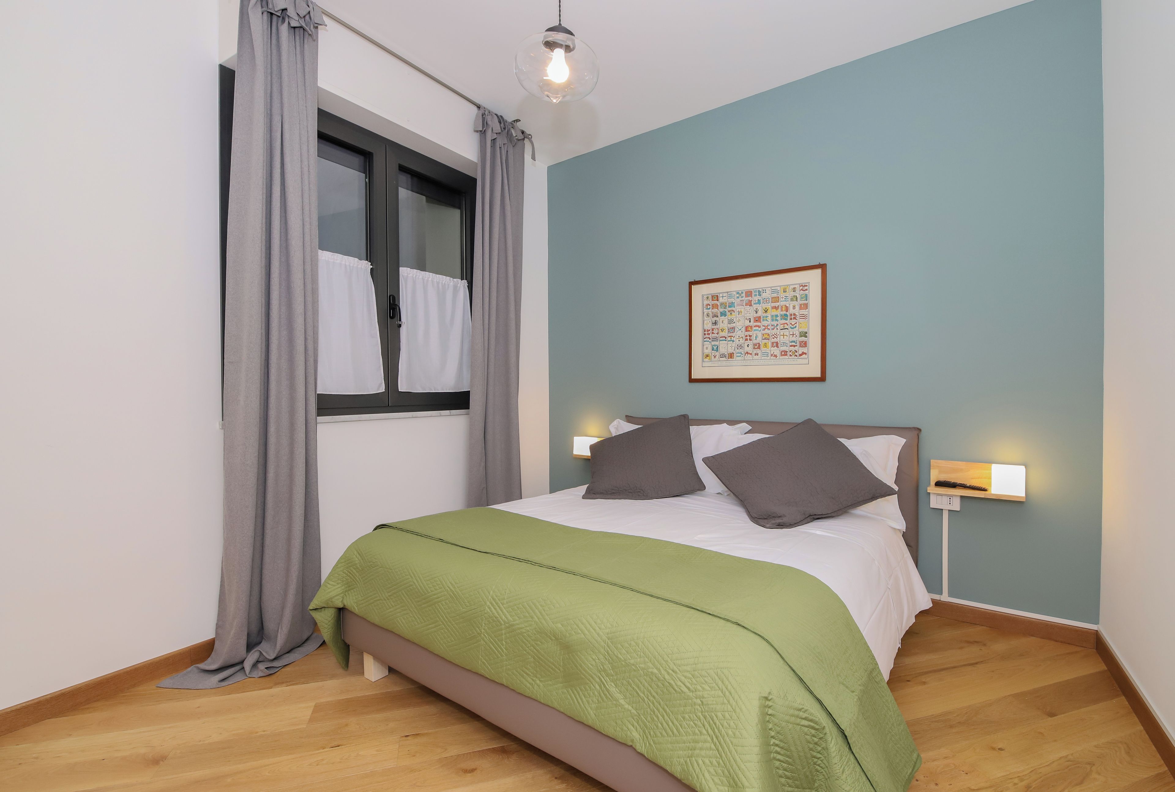 A bedroom with a bed, a window with curtains, a wall-mounted light, and a framed picture.