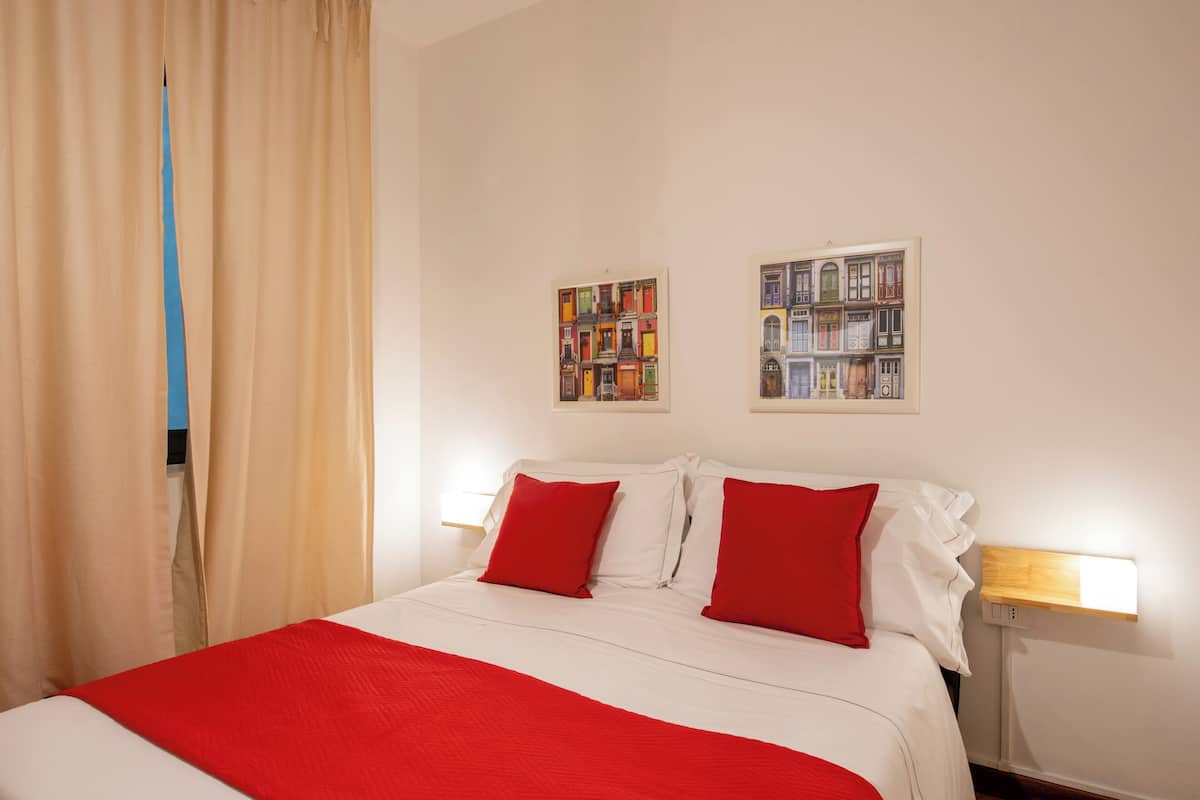 Habitación de hotel con cama, almohadas rojas, una colcha roja, dos obras de arte en la pared y una mesita de noche con lámpara.